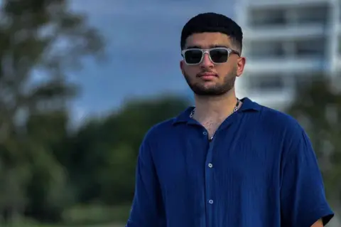 Family of Sorena Golgun A young man stands outdoors near a metal railing, wearing dark sunglasses with light-coloured frames, a smart dark-blue short-sleeved button-up shirt, black jeans, a watch on his left wrist, and a chain necklace. He has a short, black beard, from his ears to his chin. Behind him is a grassy field with scattered green leafy trees and a tall white modern multi-story building partially visible. The sky is overcast, with a mix of blue and grey clouds.