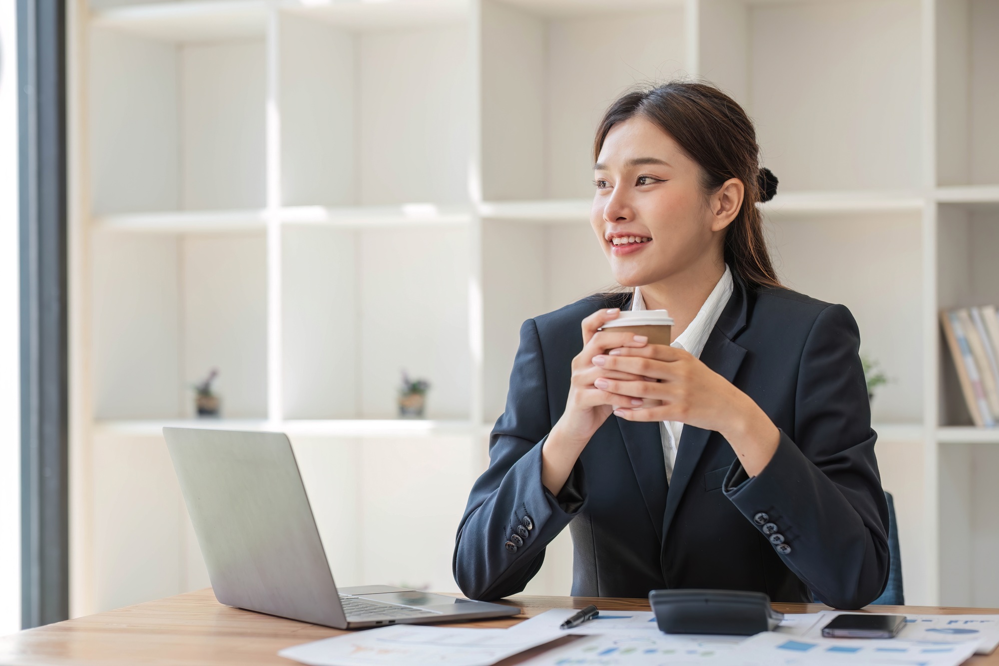 Photo of positive Asian businesswoman holds a cup of coffee imagines plan holiday in office.