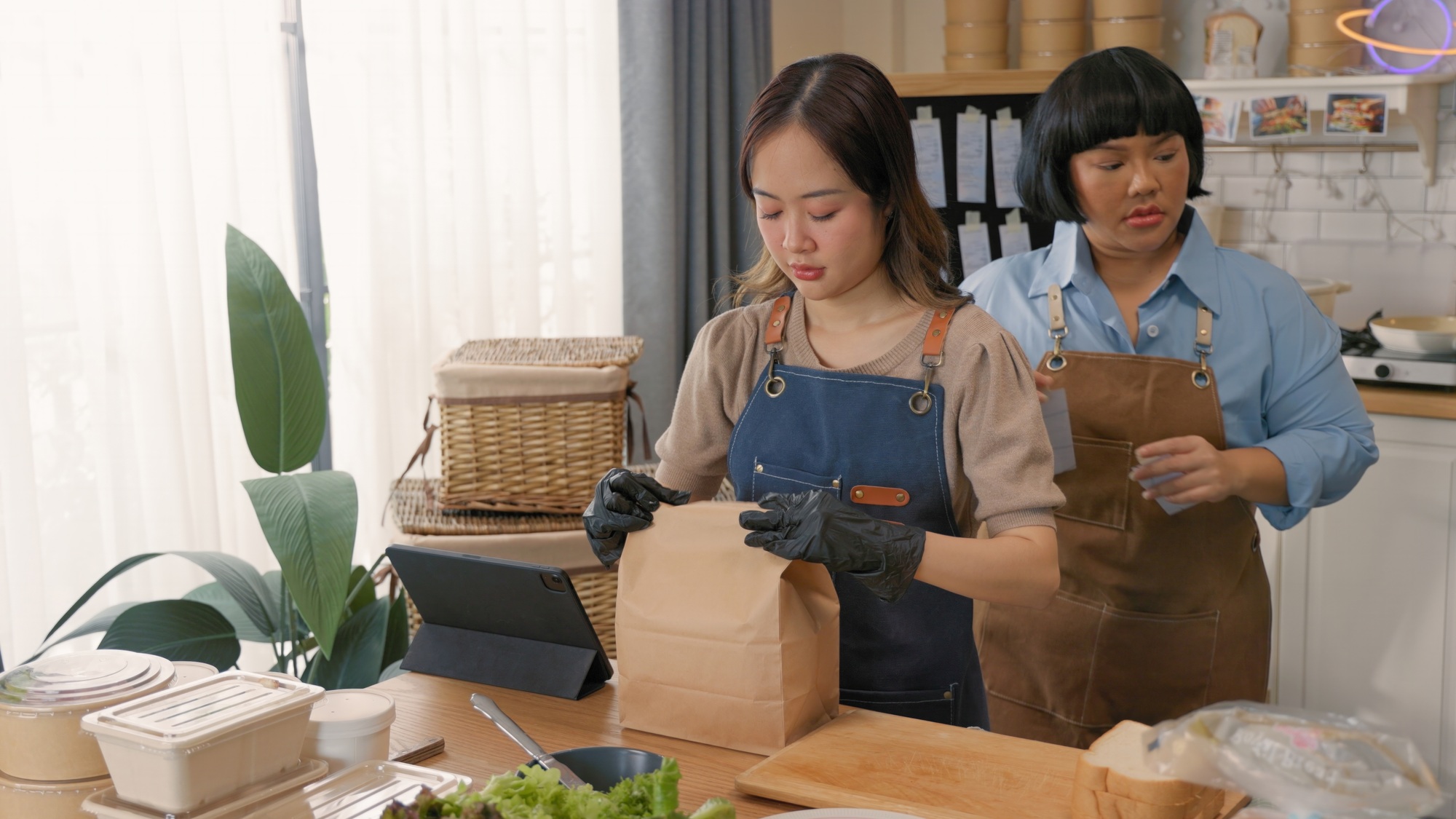 Two women working together in a kitchen, packing food into eco-friendly containers for delivery or t