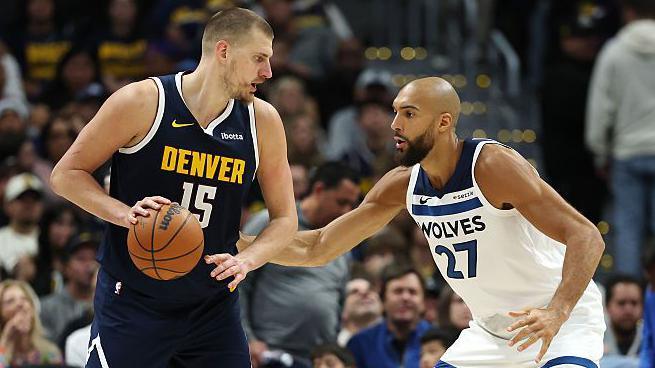 Nikola Jokic holds a basketball and stands in front of Rudy Gobert