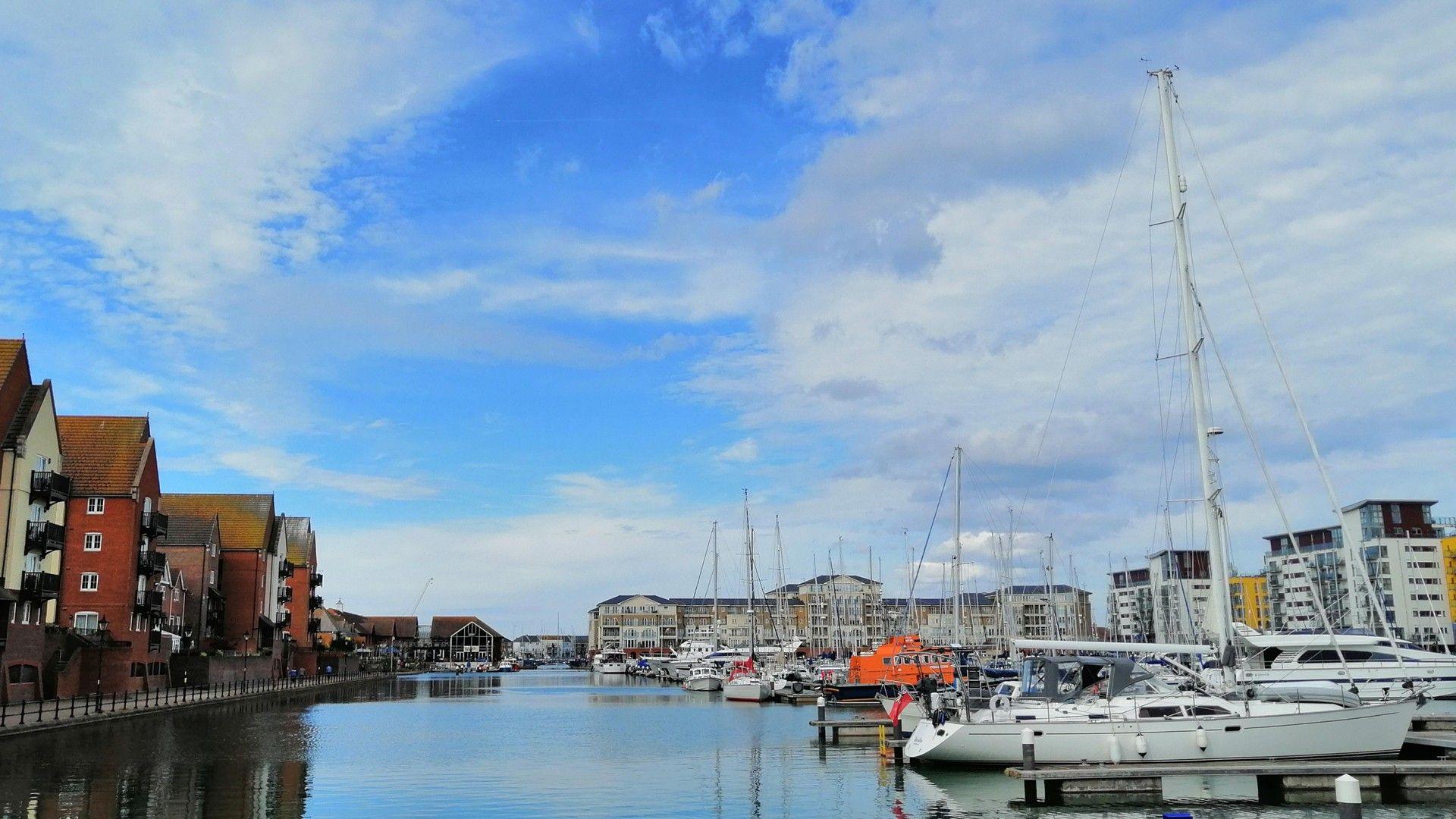 Eastbourne harbour with waterfront houses to the left and a row of moored boats on the right, on clear water and under cloudy skies
