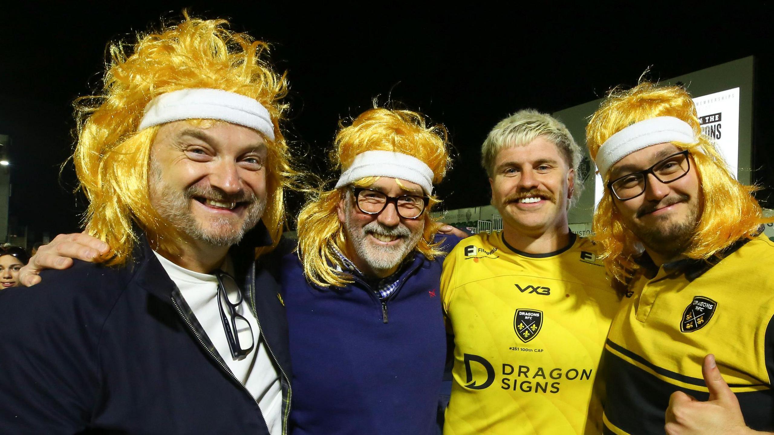 Aaron Wainwright taking a photograph with three supporters wearing blonde wigs emulating his mullet hair style.