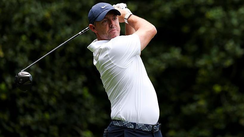 Rory McIlroy at the Truist Championship, wearing a white golf shirt and blue cap, swinging a metal wood