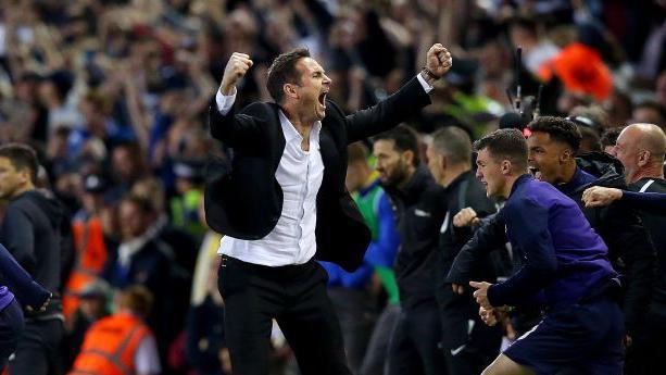 Frank Lampard celebrates on the touchline wearing a black suit and white shirt with fists clenches above his head