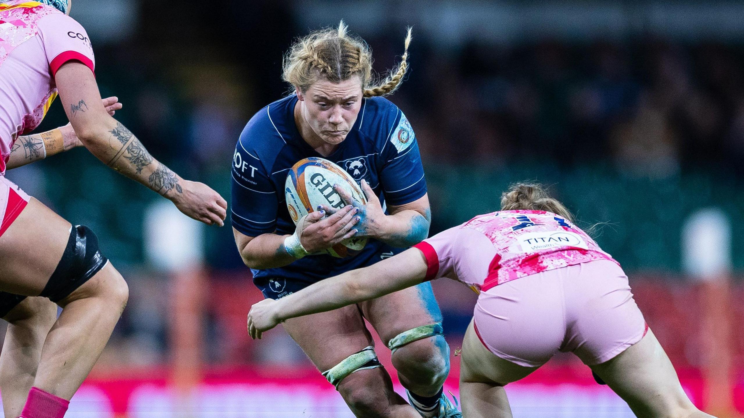 Delaney Burns runs into a tackle holding the rugby ball in two hands to her chest, as a Harlequins player bends and holds her left arm out to wrap around and tackle