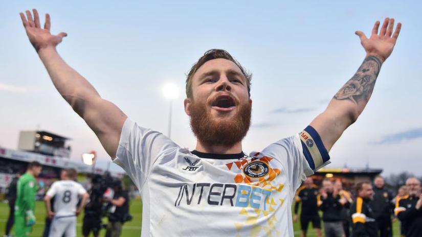Mark O'Brien in a white Newport kit with his hands in the air in celebration after a game