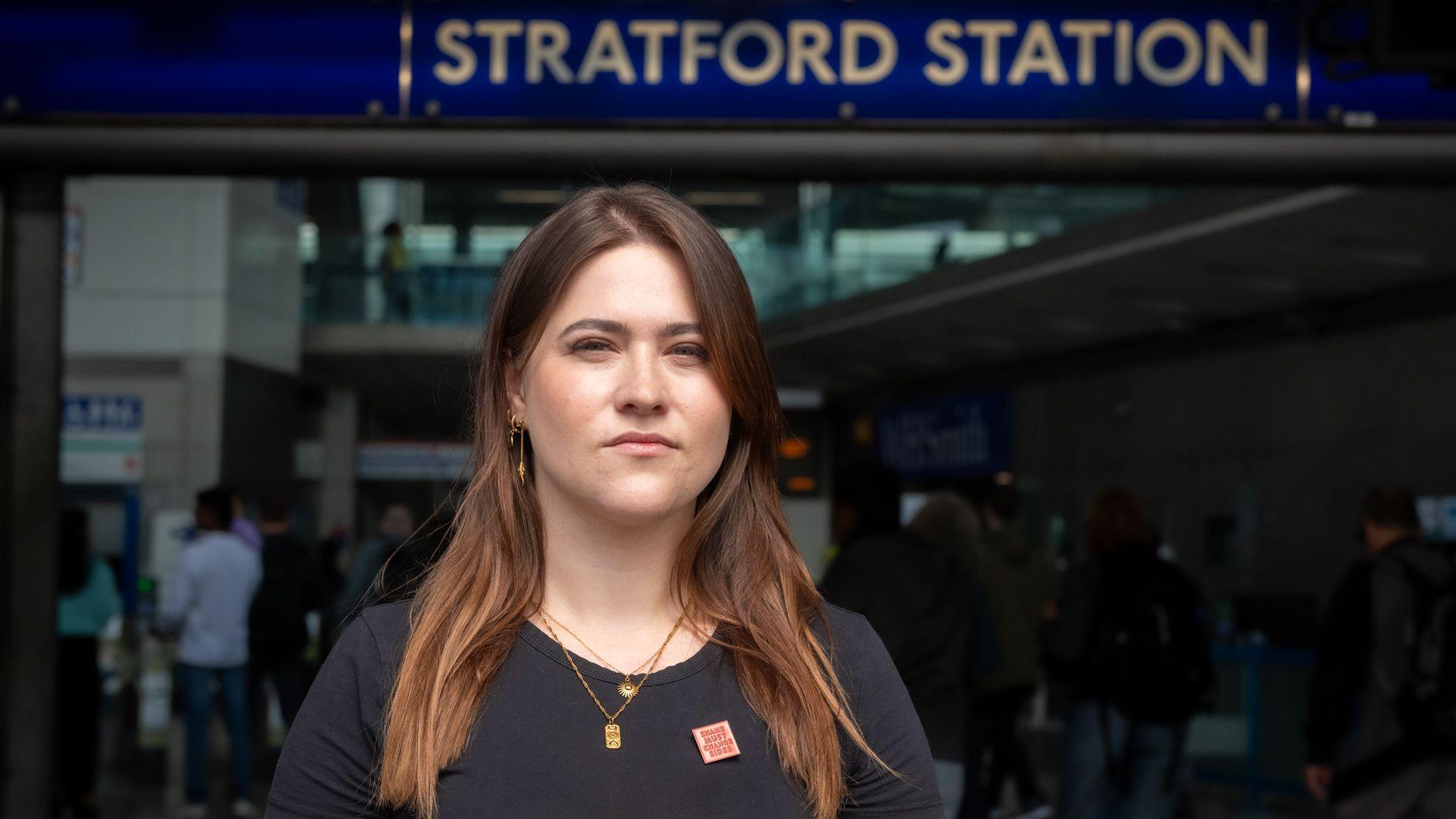 A woman in her 30s, with long brown hair in a black T-shirt is standing in front of the entrance to Stratford station. She is looking straight into the camera. She is wearing a small red badge which says 'Shame must change sides'.