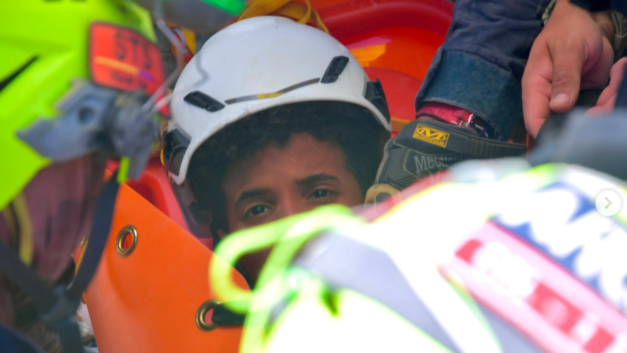 the picture shows half of the boys face - the rest is obscured by rescue workers. He's wearing a white hard hat and has dark curly hair.