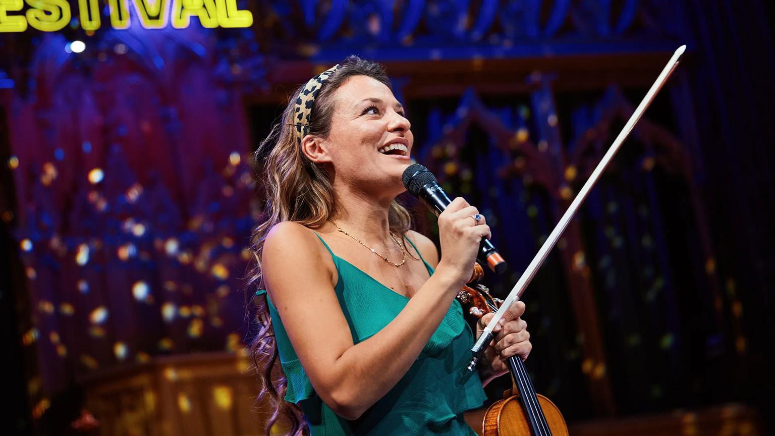 Close-up of a musician on stage at the Edinburgh International Festival holding a violin and microphone, with a bright yellow “Edinburgh International Festival” sign glowing behind and ornate architectural details lit in blue in the background.