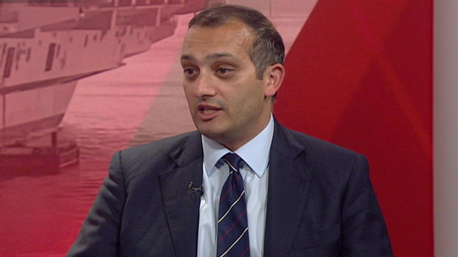 Neil Shastri-Hurst, a man with short black hair, glasses, and a dark suit and tie sits in a TV studio in front of a red tinted photo-montage background.