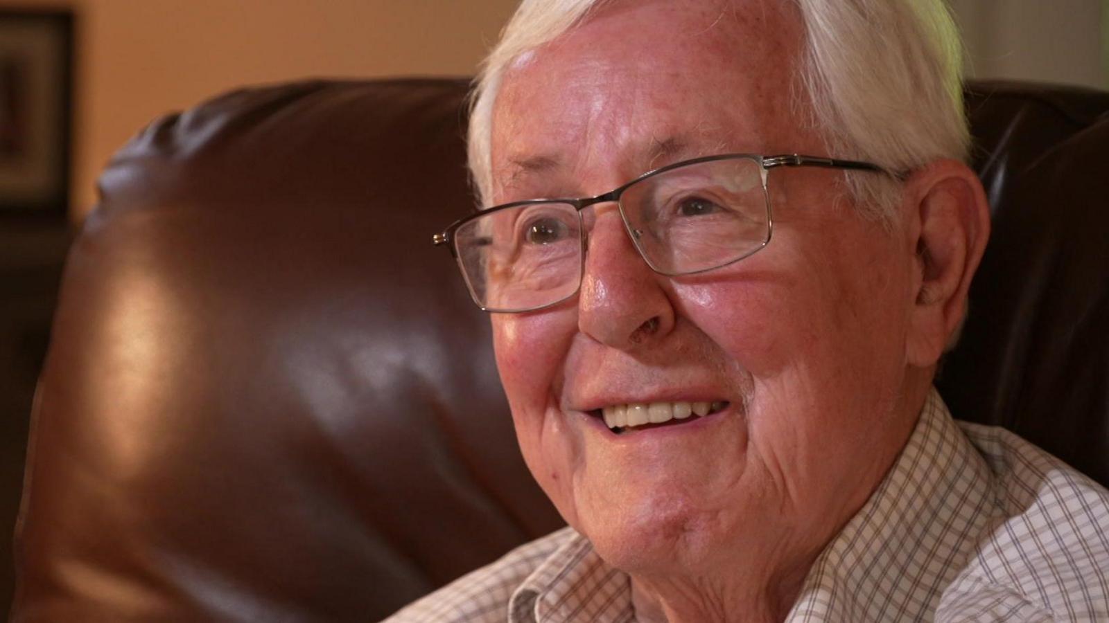 Stephen Harrington is an older man, aged 83. He has white hair and wears thin-rimmed clear-lensed glasses. He is sitting on a brown leather chair and wears a white and brown checked shirt. He is smiling.