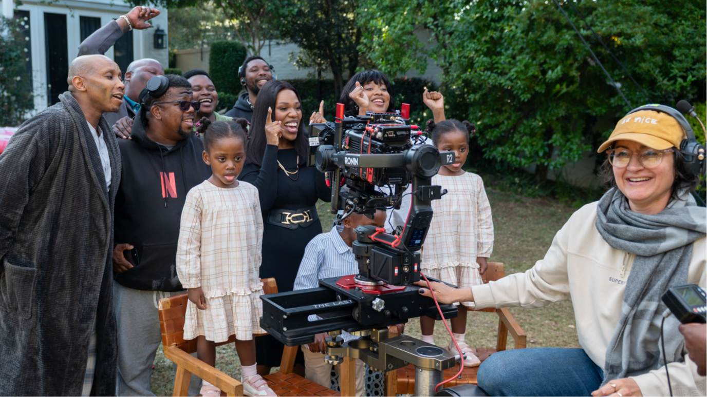 Behind the scenes at filming of The Polygamist showing some crew members and actors, including young twin girls standing on chairs in matching cheque dresses, smiling for a photo in a garden.