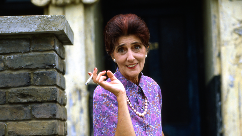 June Brown as Dot cotton in Eastenders. She is wearing a purple shirt, has short permed hair and is holding a cigarette