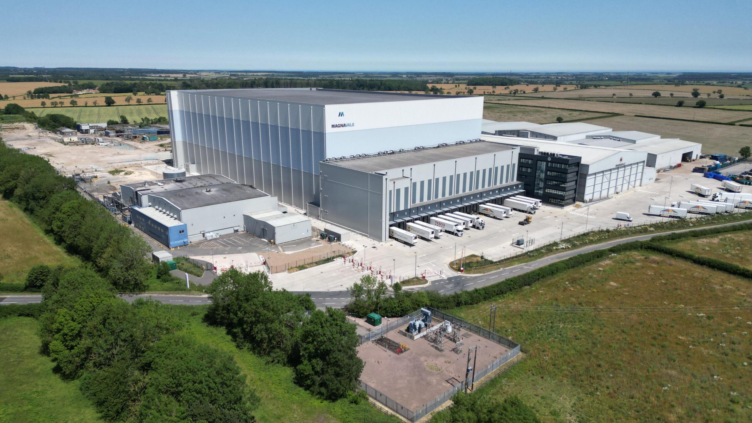 The Magnavale plant - a large warehouse building clad in white blue and grey with a distribution point at the front for HGV trailers and several smeller buildings. It is set in green fields under a blue sky.
