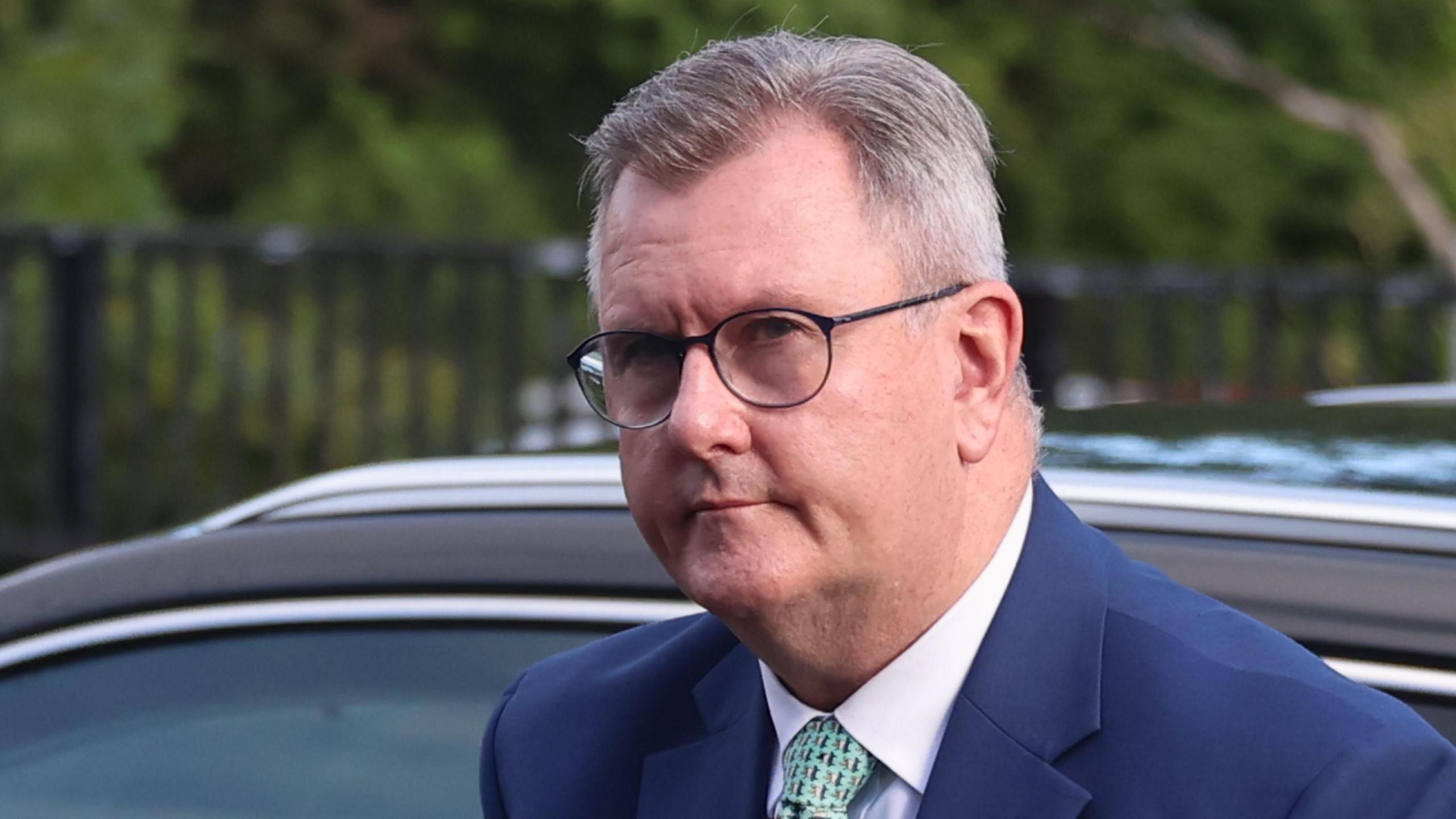 A man with short grey hair and glasses wearing a suit and tie standing outside in front of a car.
