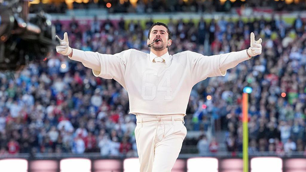 Bad Bunny performs onstage during the Apple Music Super Bowl LX Halftime Show at Levi's Stadium. He is dressed in an all-cream football jersey and trouser set.
