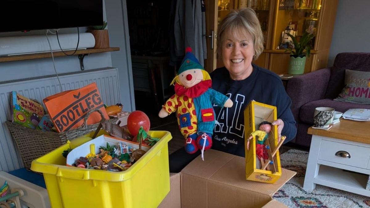 A woman with short blonde hair wearing a dark blue jumper holding up children's toys, she is surrounded by boxes of children's toys.