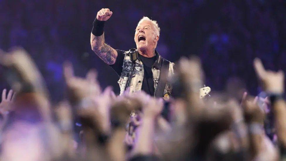 Metallica front man, James Hetfield, punches the air wearing a waistcoat, black T-shirt and wrist sweat band with short blonde hair