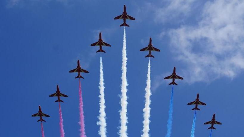 Nine red arrows are seen in formation, leaving trails of red, white and blue vapour, during a display in the sky