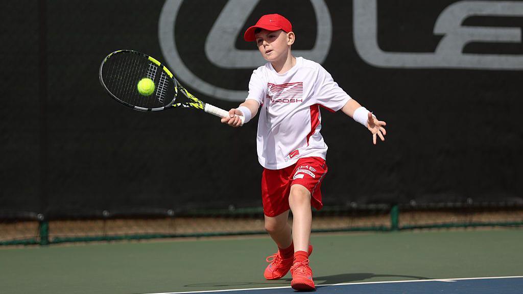 A boy is playing tennis on an outdoor court, holding a racket and striking a bright yellow ball.  The person is wearing a red cap, a white sports shirt, red shorts, and red athletic shoes.  A dark backdrop with large lettering is visible behind the court, highlighting the action in the foreground.