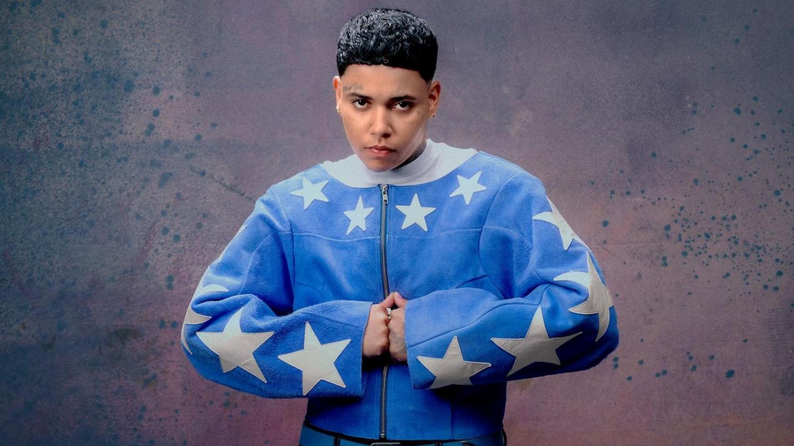 The musician KWN is pictured in a "strongman" pose, with her hands pressed together in fists. She is wearing a blue zip-up jacket with large white stars on the sleeves and collar.