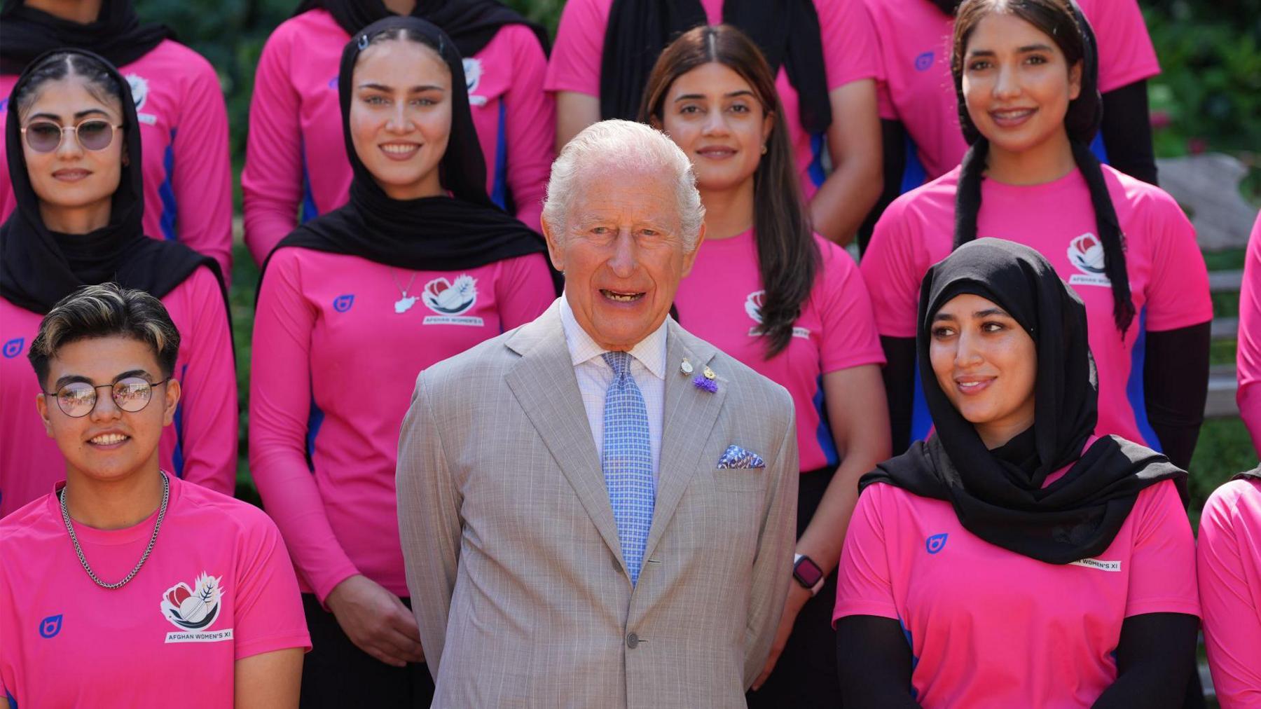 King Charles wearing a pale grey suit with white shirt and blue tie, with members of the Afghan women's cricket team - wearing hot pink T-shirts - on either side and behind him. All are looking ahead and smiling
