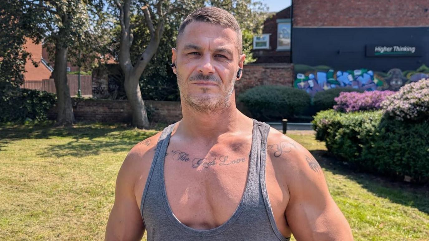 Joe, a muscular man with tattoos and salt and pepper hair, stands in front of some residential parkland with trees and flower bushes and brick walls behind him.