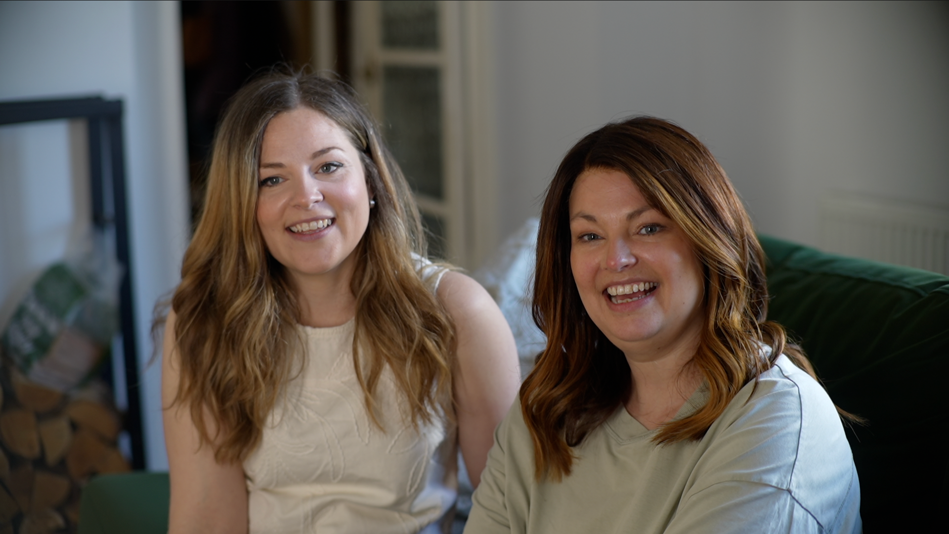 Liv on the left is wearing a white blouse, and has muddy blonde hair. She is smiling next to her older sister Lucy, She is wearing a white top, also smiling. She has brown hair and they are sat together on a sofa.