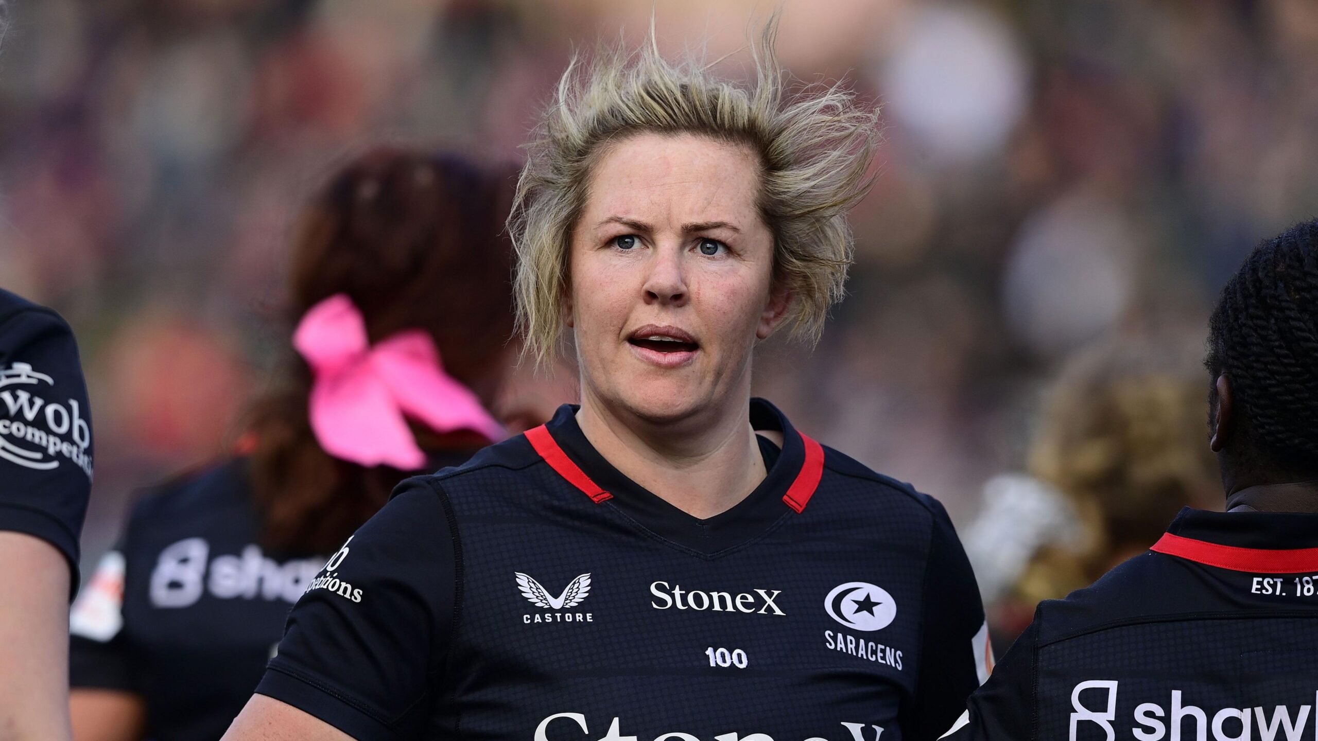 Marlie Packer looking on, wearing Saracens home dark blue kit with hair blowing in the wind. Her teammates are in the background.