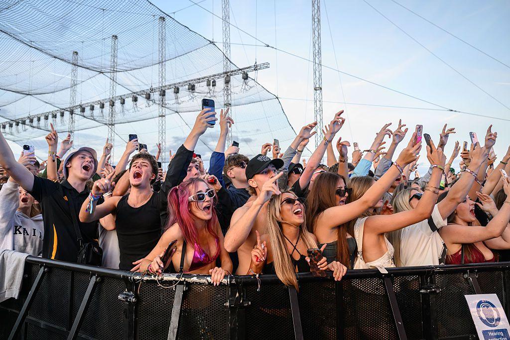Festivalgoers attend Leeds Festival at Bramham Park. They are shown with their hands in the air cheering and smiling, at the barriers in a performance.