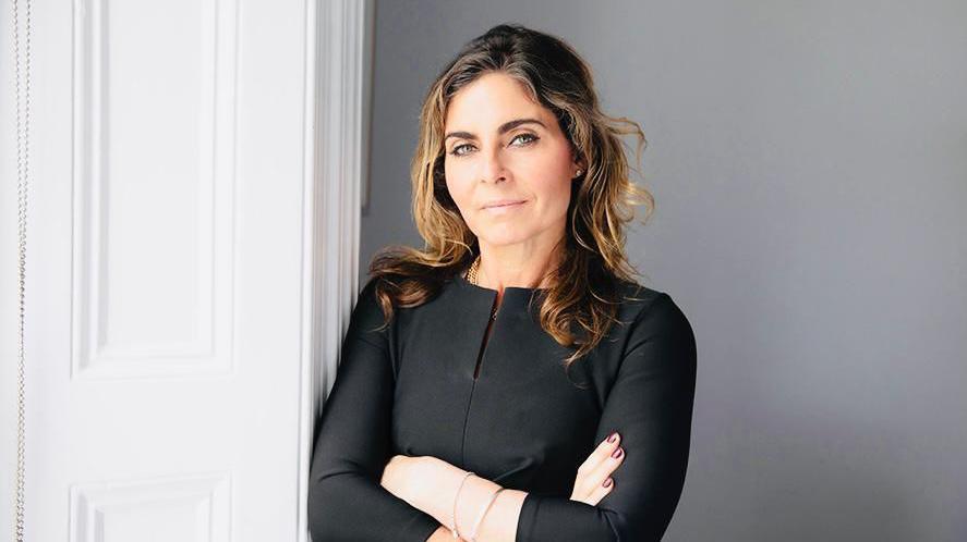 A woman with long, wavy brown hair, wearing a smart black top. She is leaning against a white door with her arms folded and and is looking at the camera with a serious expression.