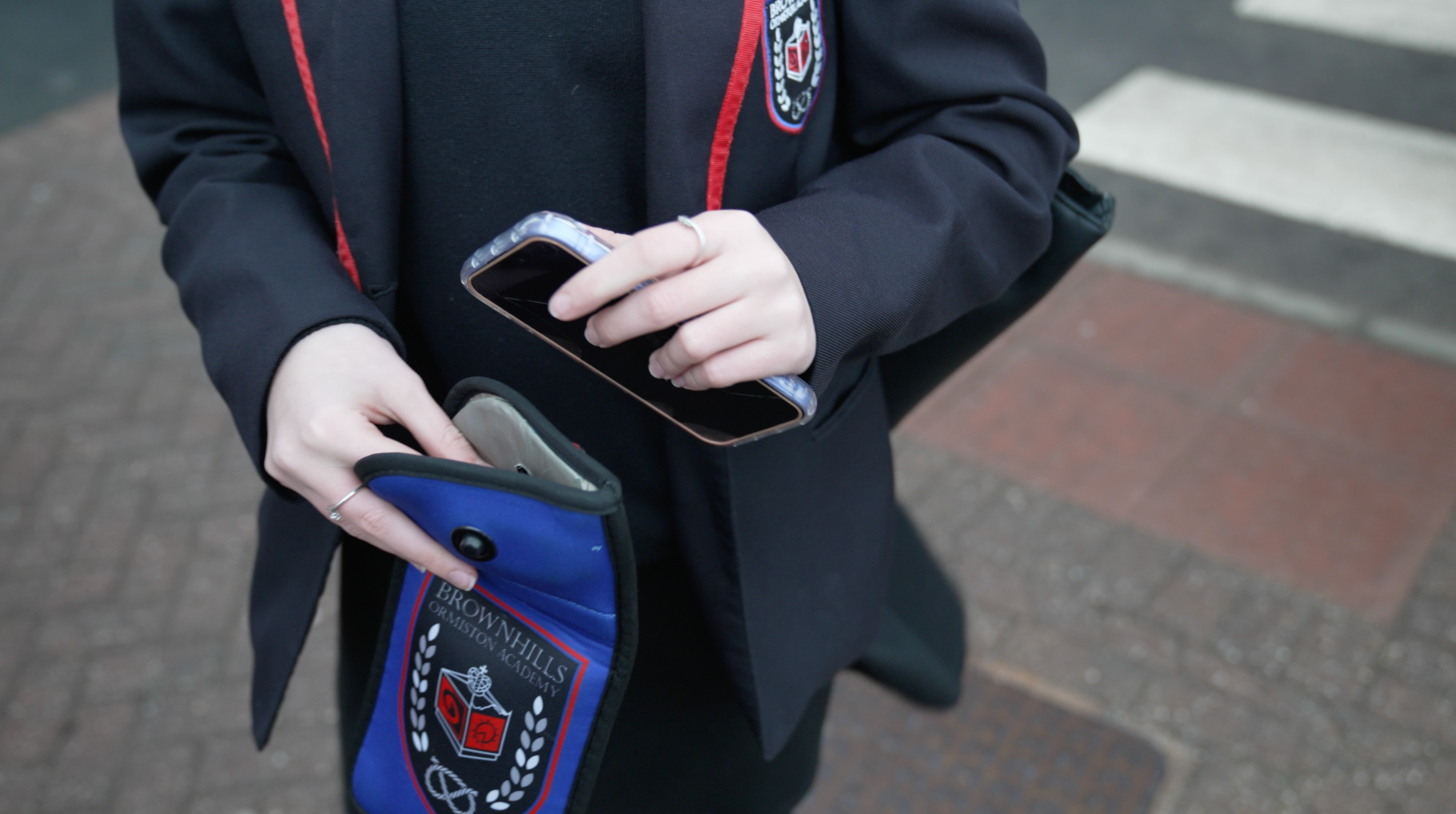 Close-up of a pupil in a dark school blazer with red trim placing a smartphone into a small blue pouch bearing the school crest, standing on a pavement.