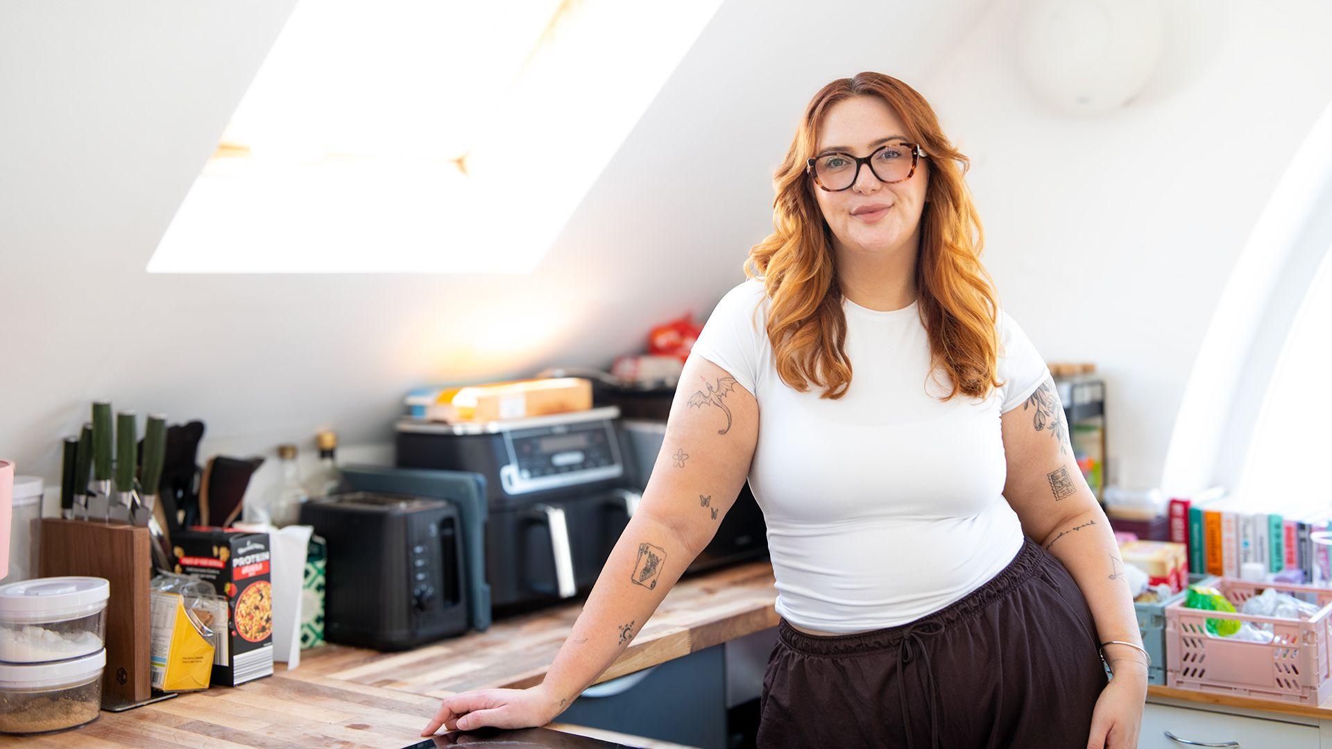 Jess King, a woman with long ginger hair and glasses, stands in front of a kitchen counter with appliances and a knife block. She is wearing a short-sleeved white top and brown trousers and has visible black tattoos at the top of her arms.