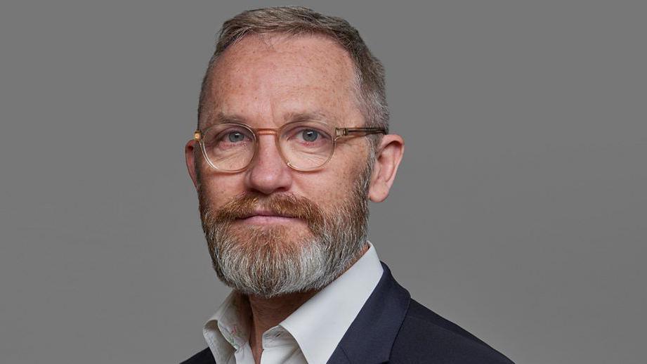 James Purnell, a middle aged man with a ginger and grey beard and glasses, poses for a formal picture
