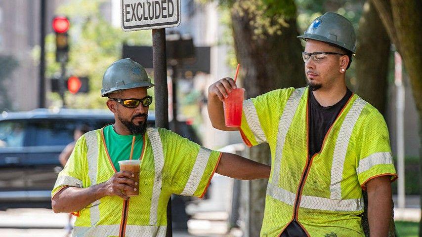 Two construction workers wearing hi-vis jackets and helmets take a drinks break in Everett, Massachusetts.