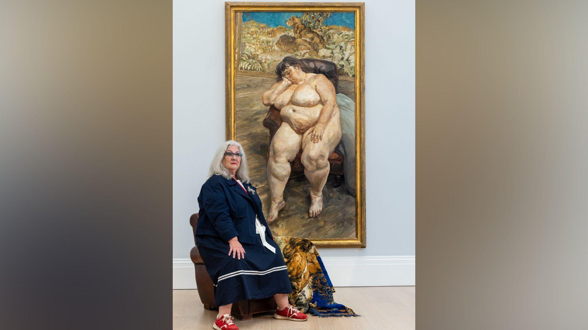 Sue Tilley is sitting on a chair in a gallery beside a large framed painting of a reclining nude figure. The gallery has a light grey wall, wooden floor, and a patterned blanket draped over the chair.