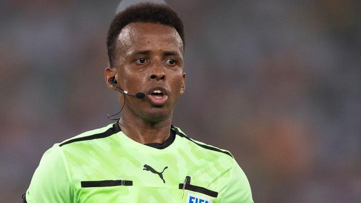 Omar Artan refereeing a match, with earpiece and microphone, and a Fifa badge on his light green shirt