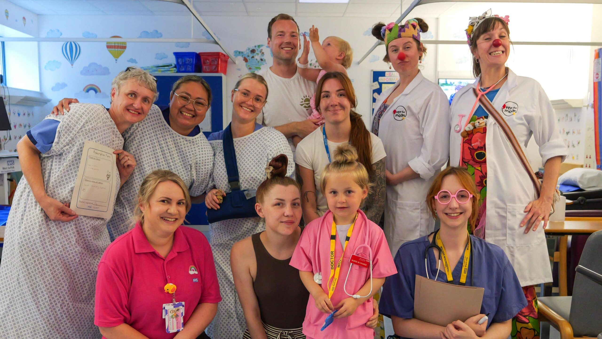A group photo of men and women - some wearing patient hospital scrubs - smiling and looking into the camera while standing in a hospital ward. In the middle at the bottom is a young girl wearing pink doctors scrubs.