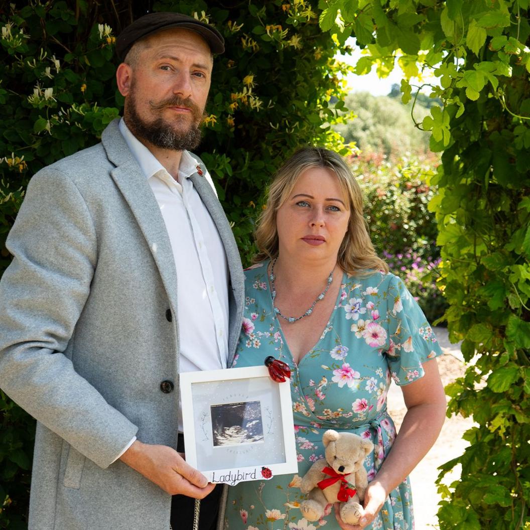 Carl Everson and Carly Wesson stand in a garden holding an ultrasound picture, in a white frame, and a teddy wearing a red ribbon.