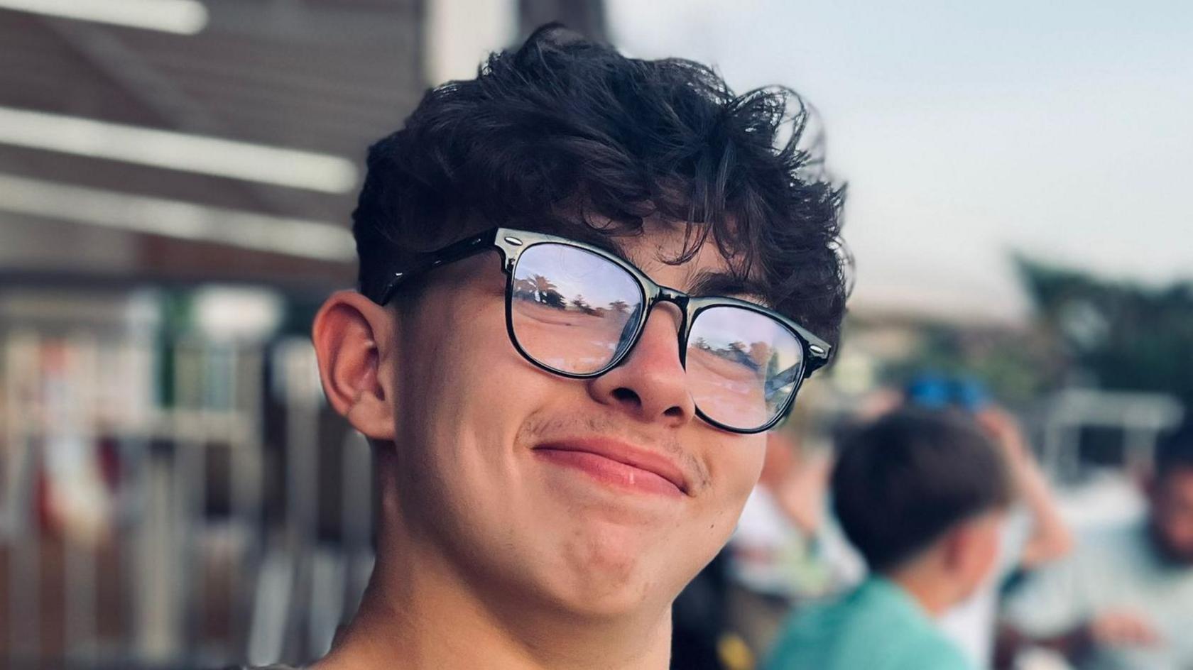 Taylor Lee, a teenage boy with dark hair and glasses with dark frames, growing the beginnings of a moustache, smiling at the camera while sitting outside at a restaurant or cafe on holiday.
