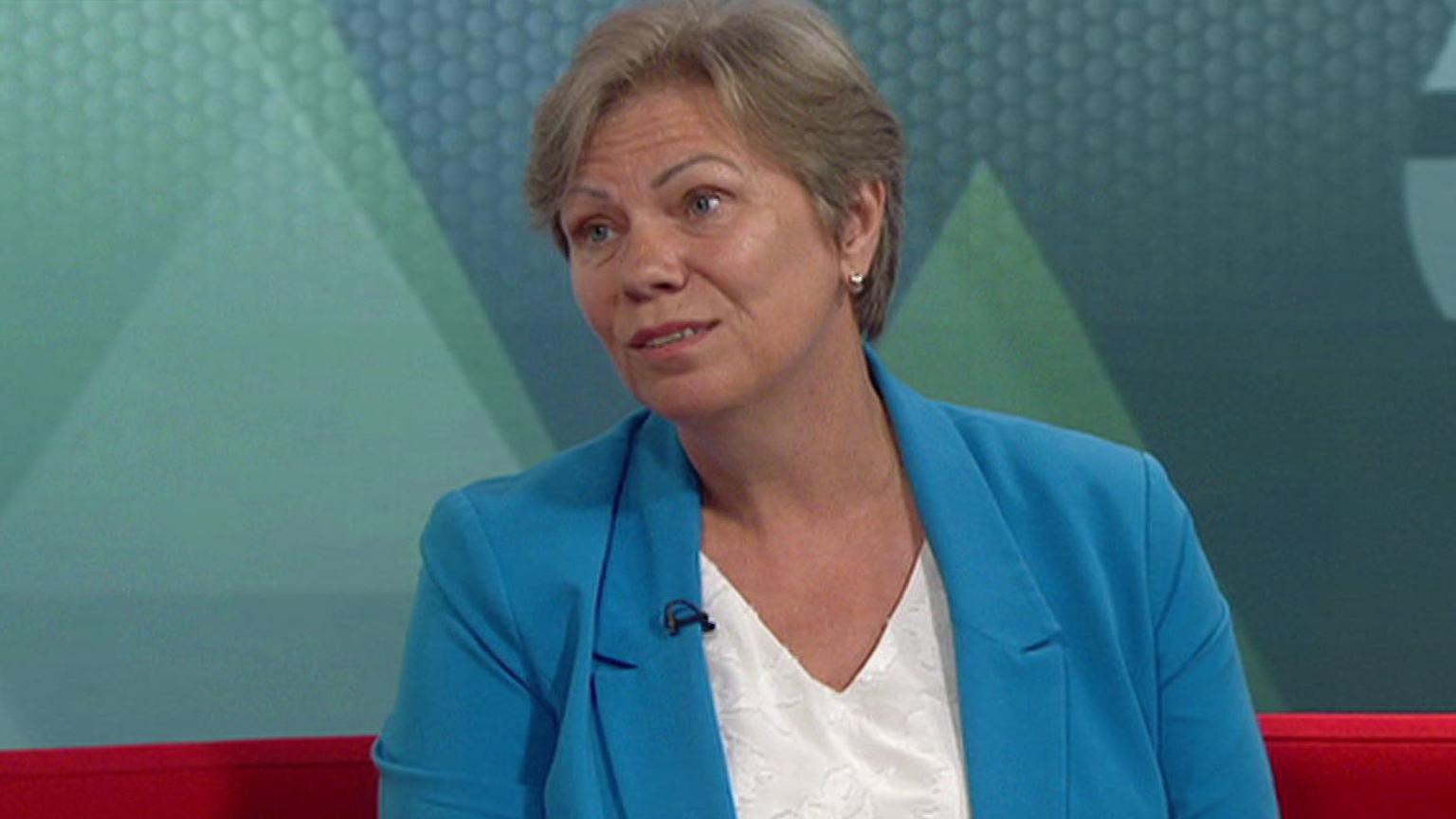 A woman with short fair hair sits on a red sofa in a TV studio, against blue and green branding on the screen behind her. She is wearing a mid-blue jacket and white top.