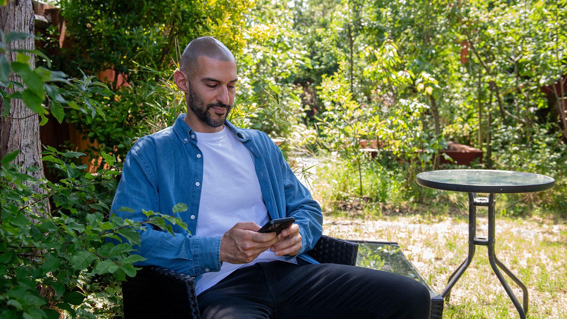 A man with a dark moustache and beard in a blue shirt, white t-shirt and back trousers, sitting in a garden, looking at his mobile phone.
