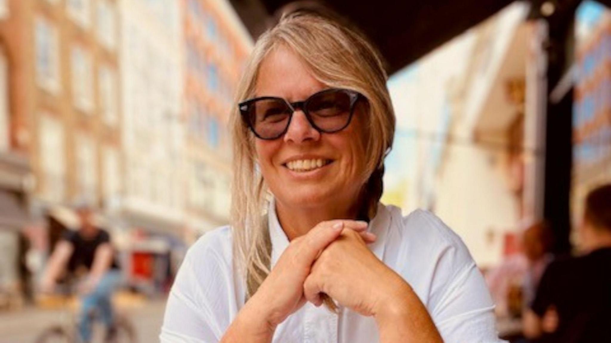 A woman with blonde hair which is tied back sits at an outdoor eatery and smiles. She wears glasses and a white blouse.