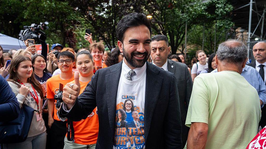 Mamdani shows a thumbs up as he smiles, surrounded by supporters.