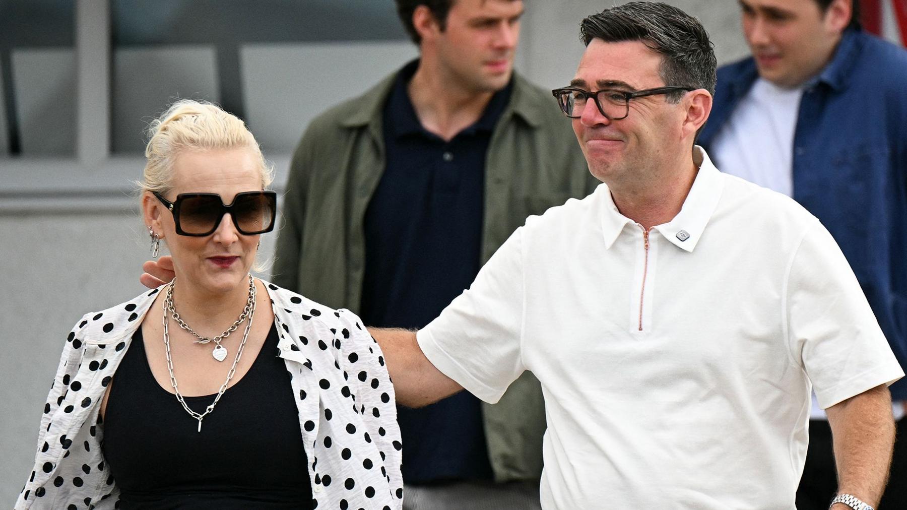 Marie-France van Heel pictured next to her husband, Andy Burnham. She is wearing a black top with a white shirt with black polka dots over the top. She has sunglasses and necklaces. Andy Burnham has his arm around her and is wearing a white polo shirt.