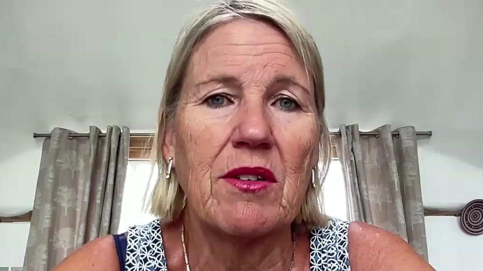 A blonde woman wearing pink lipstick looks into the camera from her home. You can see her curtains in the background.