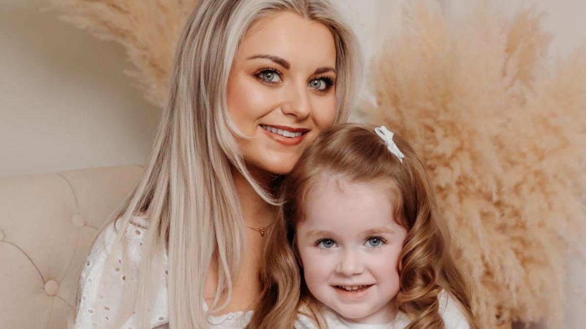 Hollie pictured on a cream chair with her mum Ann, the photo is professional. Hollie has long curly red/brown hair and is wearing a blue and white dress with a white cardigan. Ann has long blonde hair and is holding Hollie.