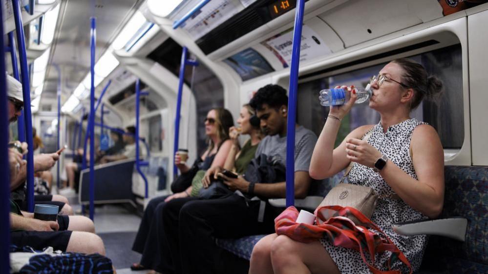 The inside of a Tube carriage with passengers sitting down. The women closest to the camera is drinking a bottle of water.