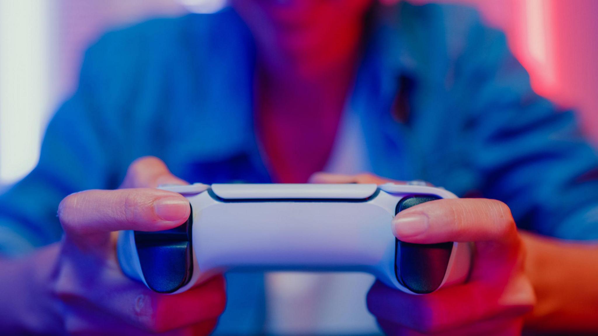 Close up of a woman's hands gripping a gaming controller, with colourful lighting reflecting on her from a monitor screen in front.