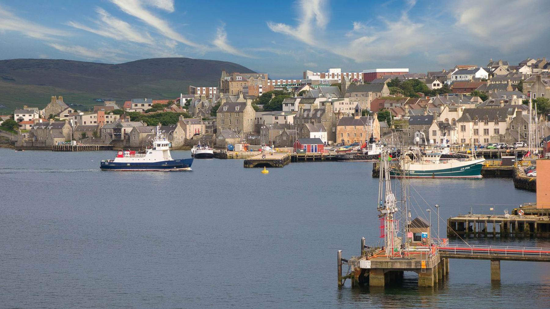 Wide view of a coastal harbour with several boats on calm water, including a ferry and fishing vessels, alongside a town of stone buildings on a gentle hillside under a blue sky with scattered clouds.